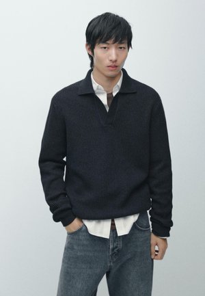 Maglione scuro a coste con collo a V, indossato sopra una camicia bianca. Indossa jeans in denim, in piedi con le mani nelle tasche contro uno sfondo chiaro.