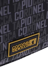 Bolsa de tela negra con un patrón repetido de texto gris y un llamativo parche de logotipo dorado y negro con el nombre de la marca.