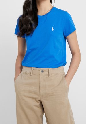 Frau trägt ein hellblaues kurzärmliges T-Shirt mit kleinem weißem Logo, in beigefarbene, hoch taillierte Hosen gesteckt, die Hände in den Taschen.