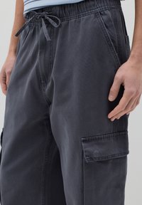 Pantaloni cargo grigi scuri con vita elastica, cordoncino e numerose tasche. Realizzati in tessuto resistente con una texture liscia.