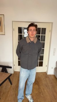 Ung mann som står inne med hendene bak på ryggen, iført en mørk jakke med beige krage, lyseblå jeans og hvite joggesko på tregulv.