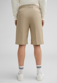 Pantaloni shorts beige con una texture liscia. Presentano una parte anteriore piatta, tasche posteriori e un taglio fino al ginocchio. Indossati con scarpe da ginnastica bianche e calzini a righe.
