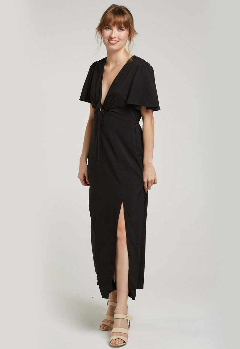 Robe maxi noire avec des manches courtes évasées, décolleté en V profond, détail noué à l'avant et fente sur le côté. Tissu lisse avec une silhouette ajustée.