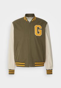 Veste bomber vert olive avec des manches crème, des finitions rayées jaunes, fermeture éclair et un écusson orange "G" sur la poitrine. Deux poches latérales.