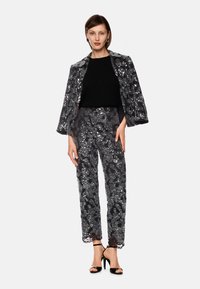 Woman wearing black top with matching sequined jacket draped on shoulders and high-waisted wide-leg sequined pants with black heels.