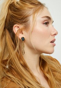 Les boucles d'oreilles pendantes en or comportent une pierre noire ovale en haut, avec des chaînes allongées en dessous, présentant une finition brillante et un design complexe.