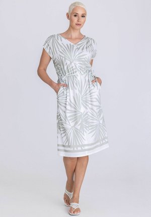 Femme aux cheveux blonds courts portant une robe blanche jusqu'aux genoux avec des motifs de feuilles grises, debout les mains dans les poches et des sandales blanches.