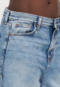 Svetlomodré denim džínsy s vysokým pásom, so zlatými nitmi a šitím. Dizajn zahŕňa predné vrecko a zips.