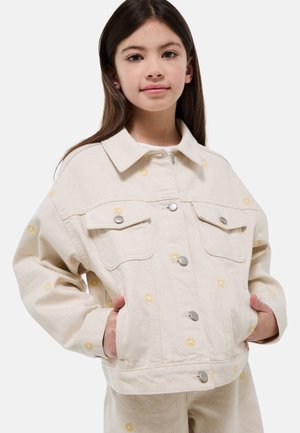Jeune fille portant une veste crème et un pantalon avec de petits motifs de fleurs jaunes, les mains dans les poches de la veste, regardant vers l'avant.