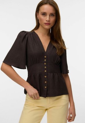 Kvinne iført en mørk brun V-halsbluse med knapper og flagrende ermer, kombinert med lysegule bukser, poserer med én hånd på hoften.