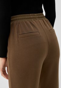 Bruine, elastische broek met een soepele textuur. Bevat twee achterzakken en heeft een relaxte pasvorm, gecombineerd met een zwart geribbeld topje.