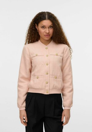 Veste bomber texturée rose clair avec un col côté, des boutons dorés et deux poches avant. Associée à un pantalon plissé noir.
