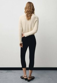 Crème, fluffy trui met lange mouwen, gecombineerd met zwarte cropped leggings. Model draagt zwarte sandalen; gezien vanuit de rug in een minimalistische setting.