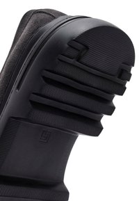 Zwarte rubberen sneakerzool met geribbelde textuur en variërende dikte, met een gladde bovenrand en een merkembleem aan de zijkant.