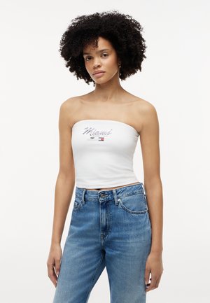 Mladá žena s kučeravými vlasmi má na sebe biely top bez ramienok s nápisom "Miami 2023" a modré džínsy s vysokým pásom na jednoduchom pozadí.