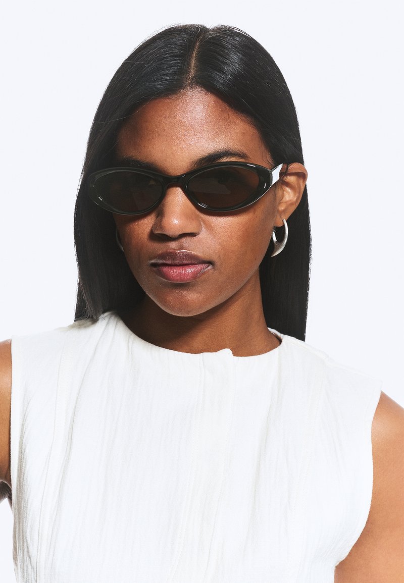 Femme aux longs cheveux raides noirs portant des lunettes de soleil noires étroites, des boucles d'oreilles argentées en anneau et un haut blanc sans manches sur fond blanc.