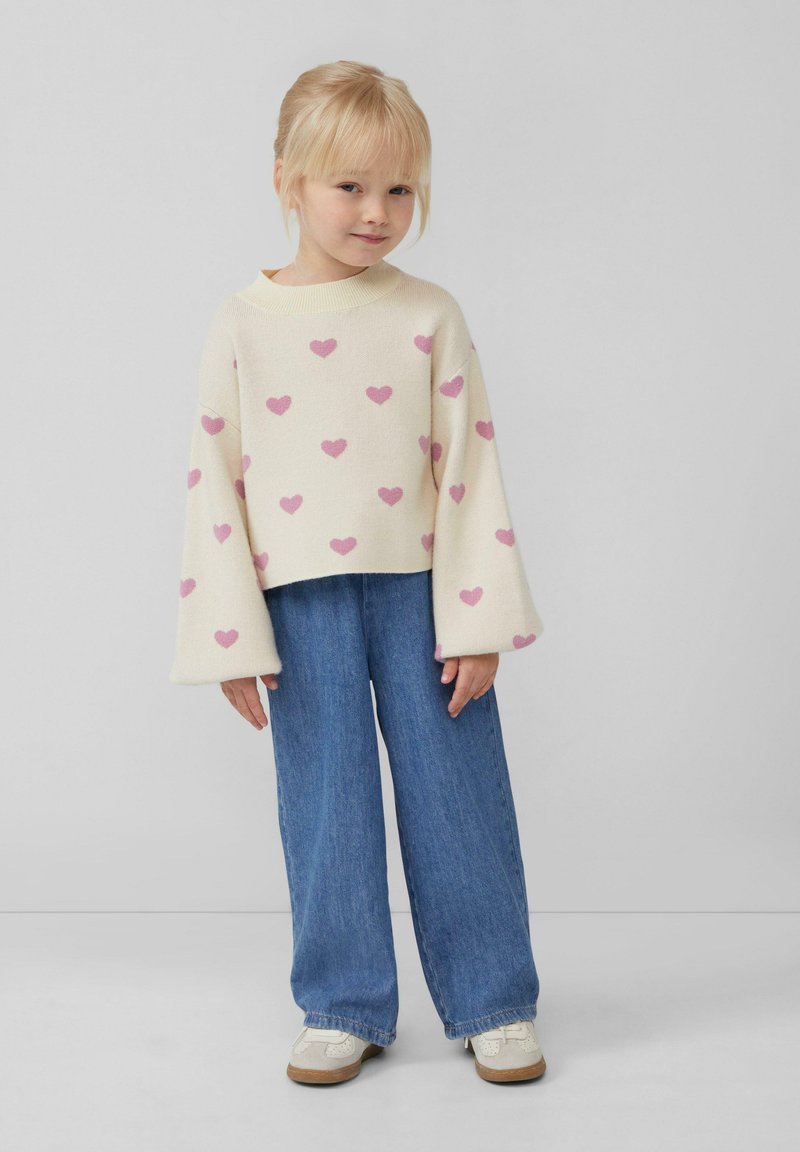 Crème trui met roze hartpatronen, brede mouwen, gecombineerd met blauwe wijde jeans en witte schoenen. Gladde textuur, casual ontwerp.