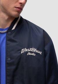 Granatowa kurtka bombowa z prążkowanym kołnierzem. Posiada haftowane logo "WrstBhn Studio" w kolorze białym. Gładka w dotyku, z ciemnoniebieskimi akcentami.