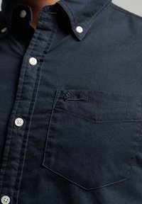 Navy blue button-up shirt made of cotton with a soft texture, featuring white buttons and a chest pocket with a branded logo.