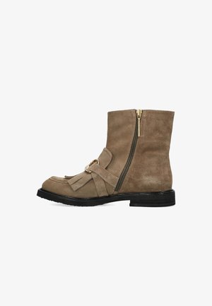 Bottine taupe en suede avec une fermeture éclair sur le côté, semelle noire plate, un accent avec un pompon et un anneau à l'avant, et des coutures décoratives sur la pointe.