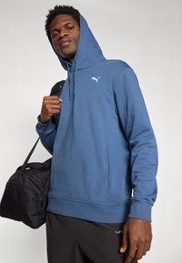 Sudadera azul con capucha y bolsillo canguro, con puños acanalados, que presenta un logo blanco de Puma en el pecho. Bolsa de gimnasio negra sostenida en una mano.