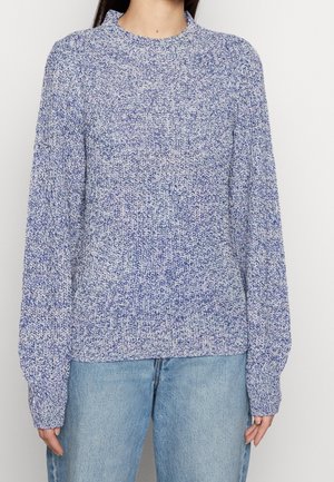 Person iført en strikket trøje med lange ærmer i blå og hvid melange kombineret med lyseblå denimjeans mod en ensfarvet baggrund.