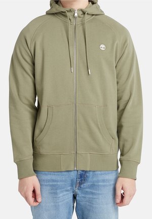 Volwassene draagt een olijfgroene zip-up hoodie met zakken aan de voorkant en een klein wit boomlogo op de linkerborst, gecombineerd met een blauwe spijkerbroek.