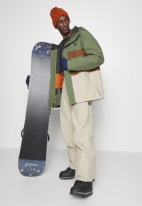 Snowboarder con una chaqueta impermeable verde y marrón, pantalones de nieve color crema, botas negras y un gorro naranja, sosteniendo una tabla de snowboard.