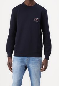 Uomo che indossa un felpa navy blue di Tommy Hilfiger con logo sul petto e jeans azzurri, in piedi davanti a uno sfondo bianco uniforme.