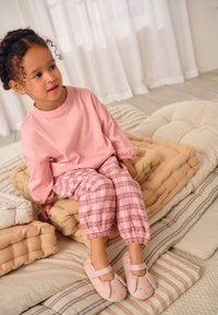 Pinkki pitkähihaispaita, punavalkoruudulliset housut joustavilla lahkeensuilla ja vaaleanpunaiset pupunkuvioiset tohvelit. Istuu kerroksittain asetetuilla ja teksturoiduilla tyynyillä.