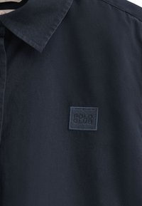 Camicia blu navy con una consistenza morbida, caratterizzata da un colletto classico e un'etichetta rettangolare ricamata sul lato sinistro.