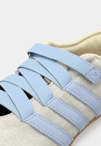 Baskets blanches avec des lacets élastiques bleu clair et trois bandes horizontales bleues. La texture semble douce avec une semelle en caoutchouc.