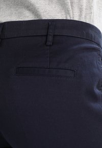 Mörkblå byxor med en slät textur, med en bakficka som har en enkel rektangulär design och en sydd söm längs kanten.