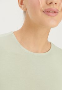 Woman wearing a light green crew-neck shirt, showing lower face with slightly parted lips and strands of blonde hair near cheek.