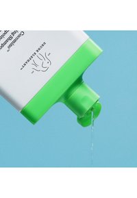 Green plastic bottle with a flip-top lid, releasing a clear liquid; white label shows text and a logo design. Background is light blue.