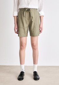 Pantalones cortos de algodón verde oliva con cinturón en la cintura, bolsillos frontales y un corte relajado. Combinados con zapatos negros y calcetines blancos.