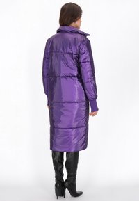 Borbottino lungo e lucido di colore viola con colletto alto, polsini a coste e cuciture orizzontali. Abbinato a stivali neri con tacco a spillo fino al ginocchio.