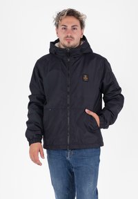 Uomo che indossa una giacca nera con cappuccio, chiusa con cerniera e con un patch del logo, jeans blu, con una mano in tasca, in piedi su uno sfondo bianco.