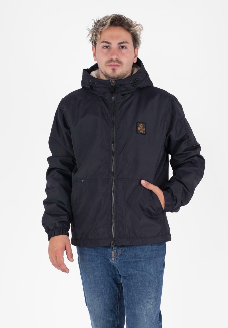 Uomo che indossa una giacca nera con cappuccio, chiusa con cerniera e con un patch del logo, jeans blu, con una mano in tasca, in piedi su uno sfondo bianco.