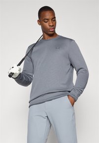 Gray long-sleeve athletic shirt with a textured fabric and subtle logo on the chest, paired with light gray pants and a golf club in hand.