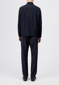 Homme portant une chemise à manches longues bleu marine foncé et un pantalon assorti, debout de dos sur un fond blanc uni.