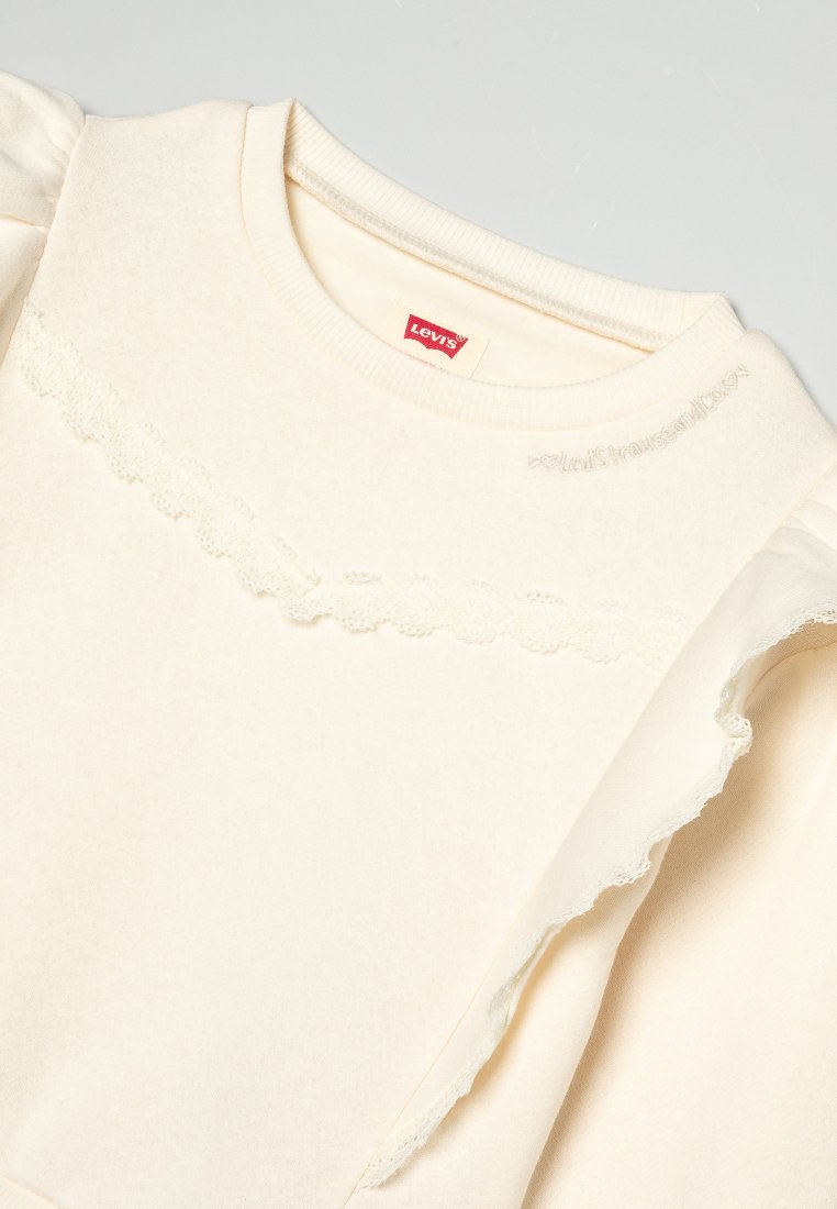 Cremfarbener Sweatshirt mit strukturierten Ärmeln und Rüschen, gewellter Spitze am Ausschnitt und gesticktem Logo in der Mitte.