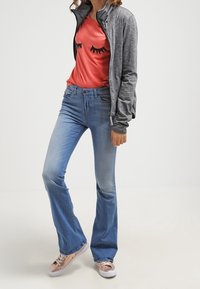 Femme portant une veste à fermeture éclair grise, un t-shirt corail avec des imprimés d'œil fermé, un jean évasé bleu clair, et des baskets en or rose.