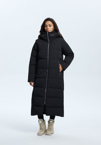 Sort lang pufferjakke med høj krave, lynlås lukning, sidelommer og quiltet tekstur. Båret over sorte leggings og brune støvler.
