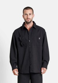 Hombre con cabello corto y barba vistiendo una camisa negra abotonada texturizada con bolsillos en el pecho, uno de los cuales sostiene unas gafas de sol negras, de pie contra un fondo blanco.