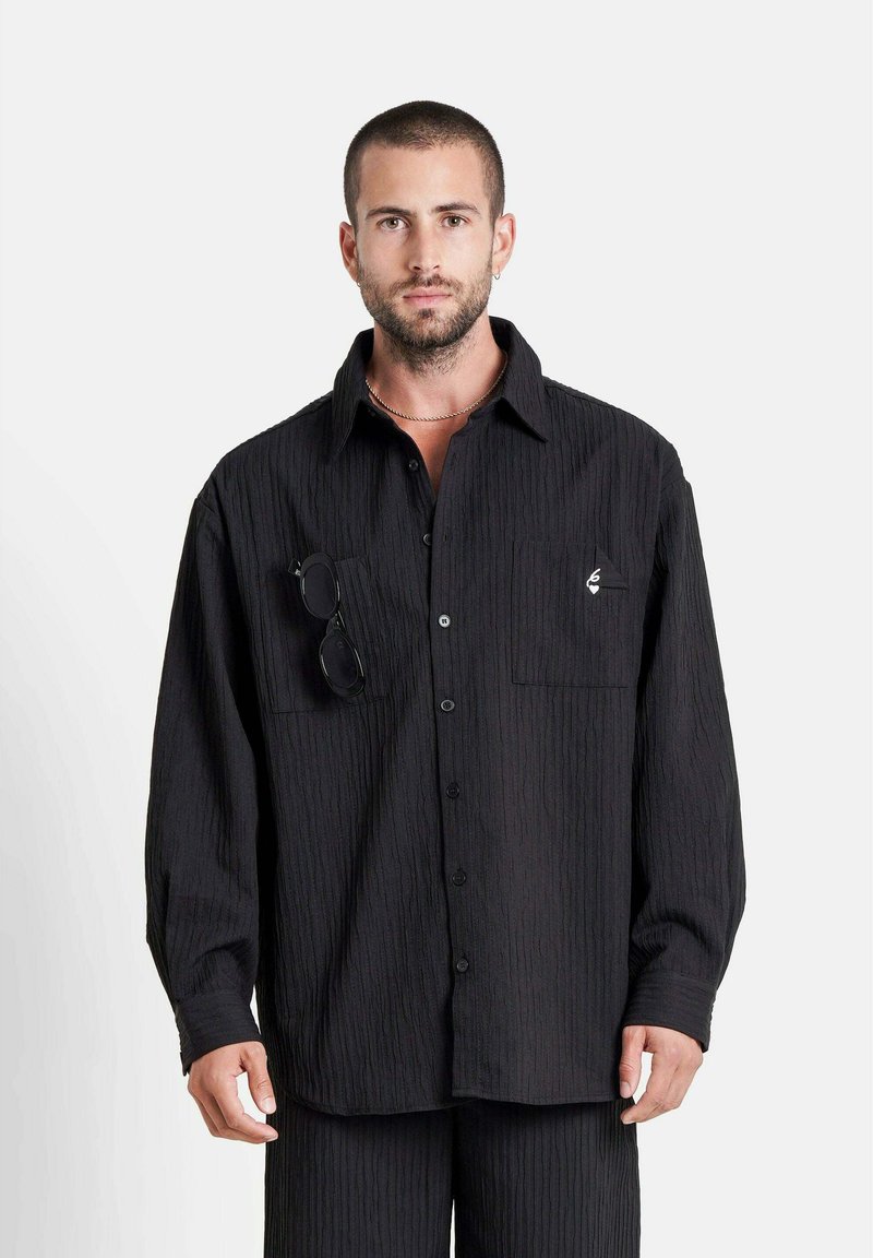 Hombre con cabello corto y barba vistiendo una camisa negra abotonada texturizada con bolsillos en el pecho, uno de los cuales sostiene unas gafas de sol negras, de pie contra un fondo blanco.