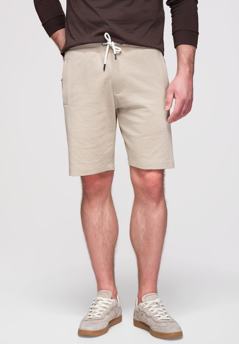 Homme portant un short beige avec cordon, des baskets claires et une chemise à manches longues foncée, debout devant un fond blanc.