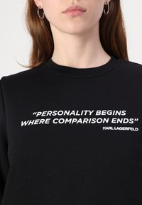 Sort sweatshirts med hvid tekst, der står "PERSONLIGHEDEN BEGYNDER HVOR SAMMENLIGNINGEN SLUTTER," samt mærkenavnet Karl Lagerfeld.
