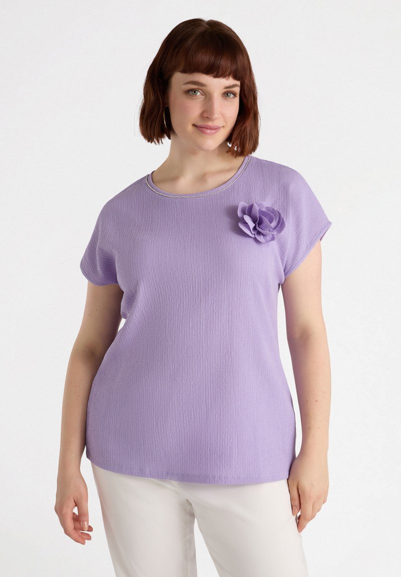 Lyhyet hihat ja tekstuuripintainen laventelinvärinen toppi, pyöreä kaula-aukko ja koristeellinen kangaskukka vasemmalla puolella. Yhdistetty valkoisiin housuihin.