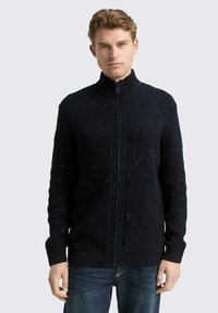 Gebreide ritstrui in donkerblauw met een gestructureerd patroon en spikkels van contrasterende kleuren. Heeft een hoge kraag en aangesloten mouwen.